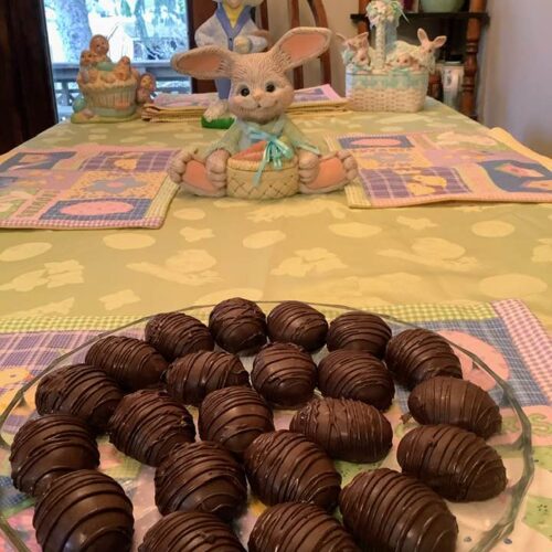 A glass plate filled with homemade chocolate-covered peanut butter eggs featuring a dark chocolate drizzle, sitting on an Easter-themed tablecloth with ceramic bunnies in the background.