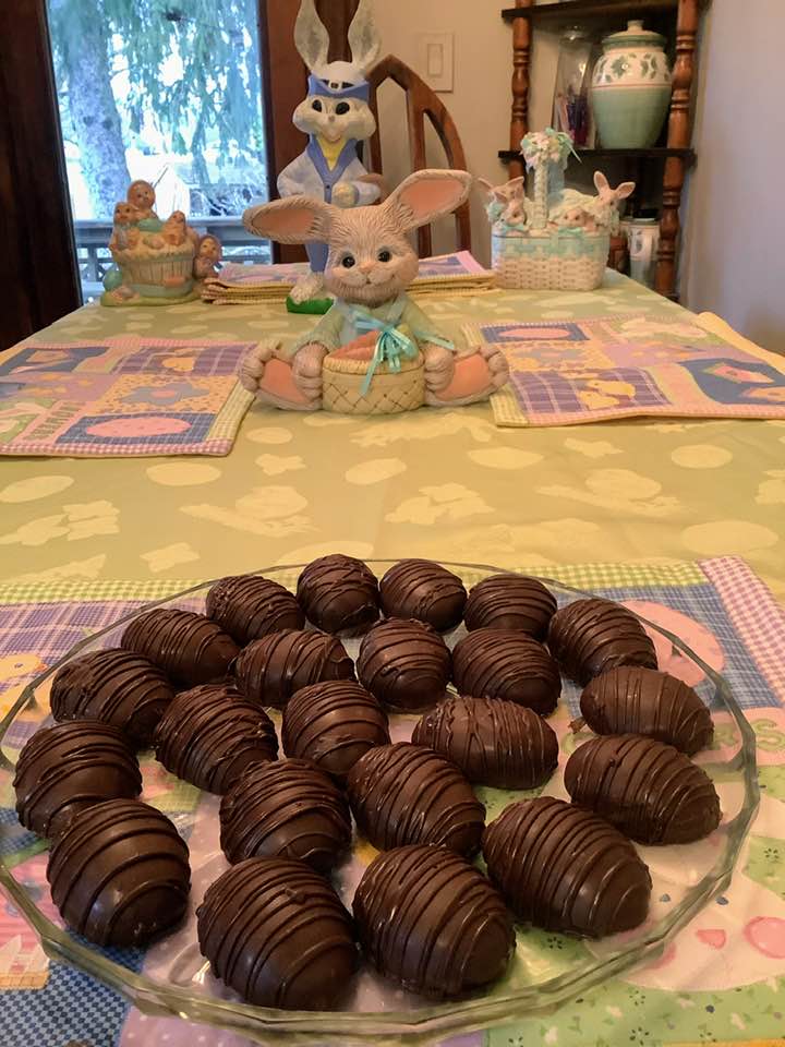 A glass plate filled with homemade chocolate-covered peanut butter eggs featuring a dark chocolate drizzle, sitting on an Easter-themed tablecloth with ceramic bunnies in the background.