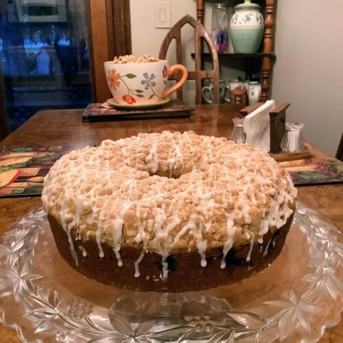 A whole Sour Cream Blueberry Coffee Cake sitting on a glass cake stand, featuring a golden crumb topping and a white glaze drizzle