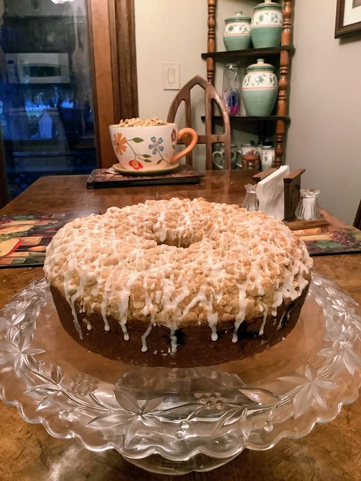 A whole Sour Cream Blueberry Coffee Cake sitting on a glass cake stand, featuring a golden crumb topping and a white glaze drizzle