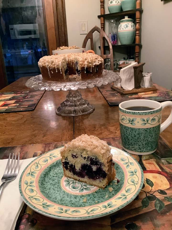 A slice of blueberry coffee cake on a floral plate next to a cup of black coffee, showing the swirl of blueberries and crumb filling in the center.
