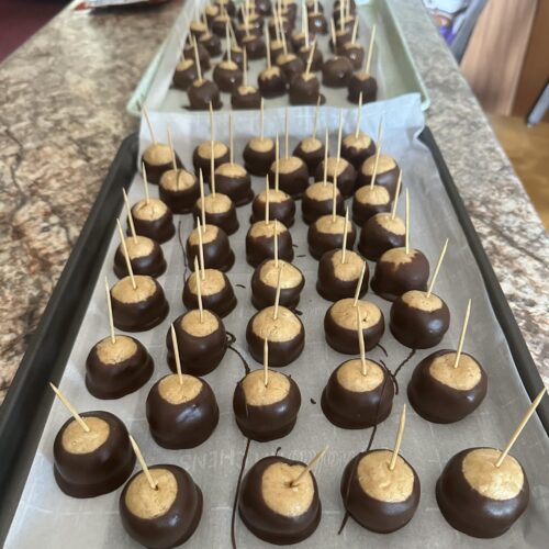 A large baking sheet lined with parchment paper filled with peanut butter balls, each with a wooden toothpick inserted, some already dipped in chocolate and some waiting to be dipped