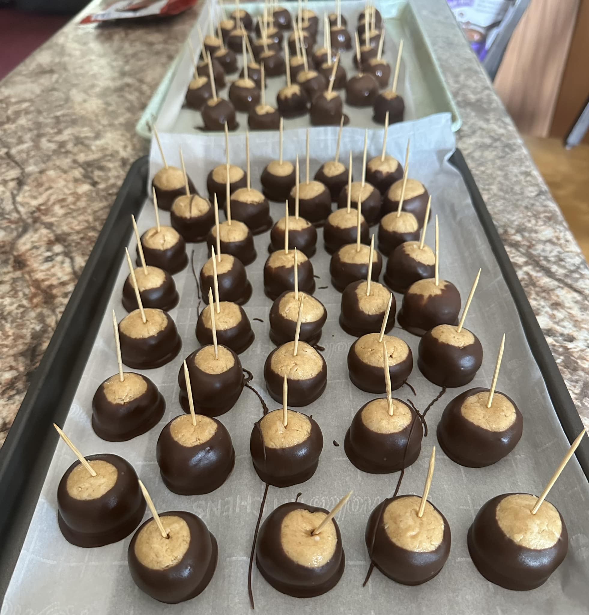A large baking sheet lined with parchment paper filled with peanut butter balls, each with a wooden toothpick inserted, some already dipped in chocolate and some waiting to be dipped