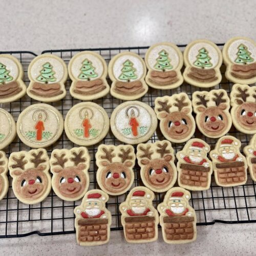 A wire rack filled with decorated Christmas cookies including smiling snowmen with red scarves, candy canes with gold bows, holly leaves with red berries, and colorful knit-texture mittens.