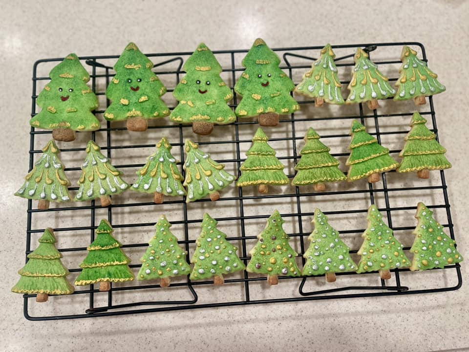 A batch of green Christmas tree cookies in various styles, some with smiley faces and others with elegant metallic gold and silver garlands piped on