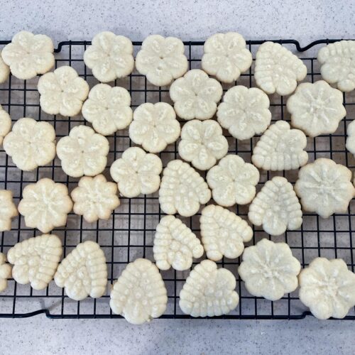 Golden baked Classic Butter Spritz Cookies in flower and leaf shapes cooling on a black wire rack
