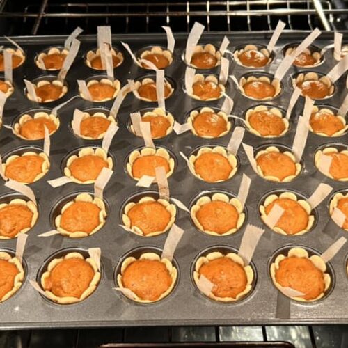 Close up of unbaked pumpkin pie tassies in a muffin tin, showing the creamy orange pumpkin filling inside the raw dough shells with parchment lifting strips visible.