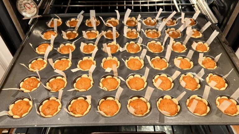 Close up of unbaked pumpkin pie tassies in a muffin tin, showing the creamy orange pumpkin filling inside the raw dough shells with parchment lifting strips visible.