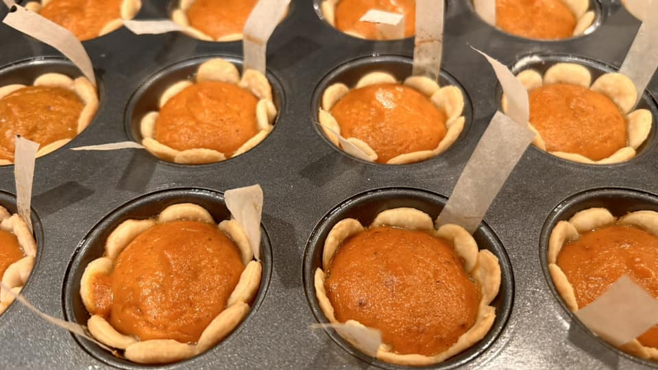 Close up of unbaked pumpkin pie tassies in a muffin tin, showing the creamy orange pumpkin filling inside the raw dough shells with parchment lifting strips visible.