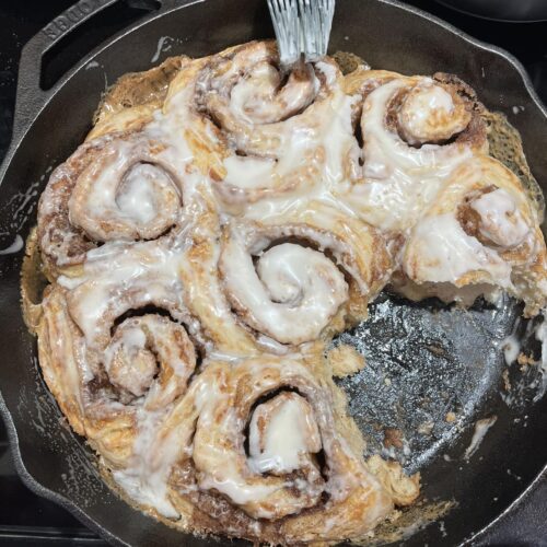 A top-down view of golden brown sourdough cinnamon rolls in a cast iron skillet being brushed with a white cream cheese glaze.