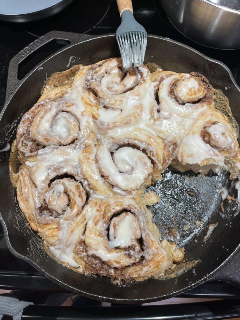 A top-down view of golden brown sourdough cinnamon rolls in a cast iron skillet being brushed with a white cream cheese glaze.