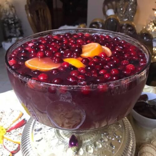 A crystal punch bowl filled with dark red Christmas punch, topped with floating orange slices and fresh cranberries