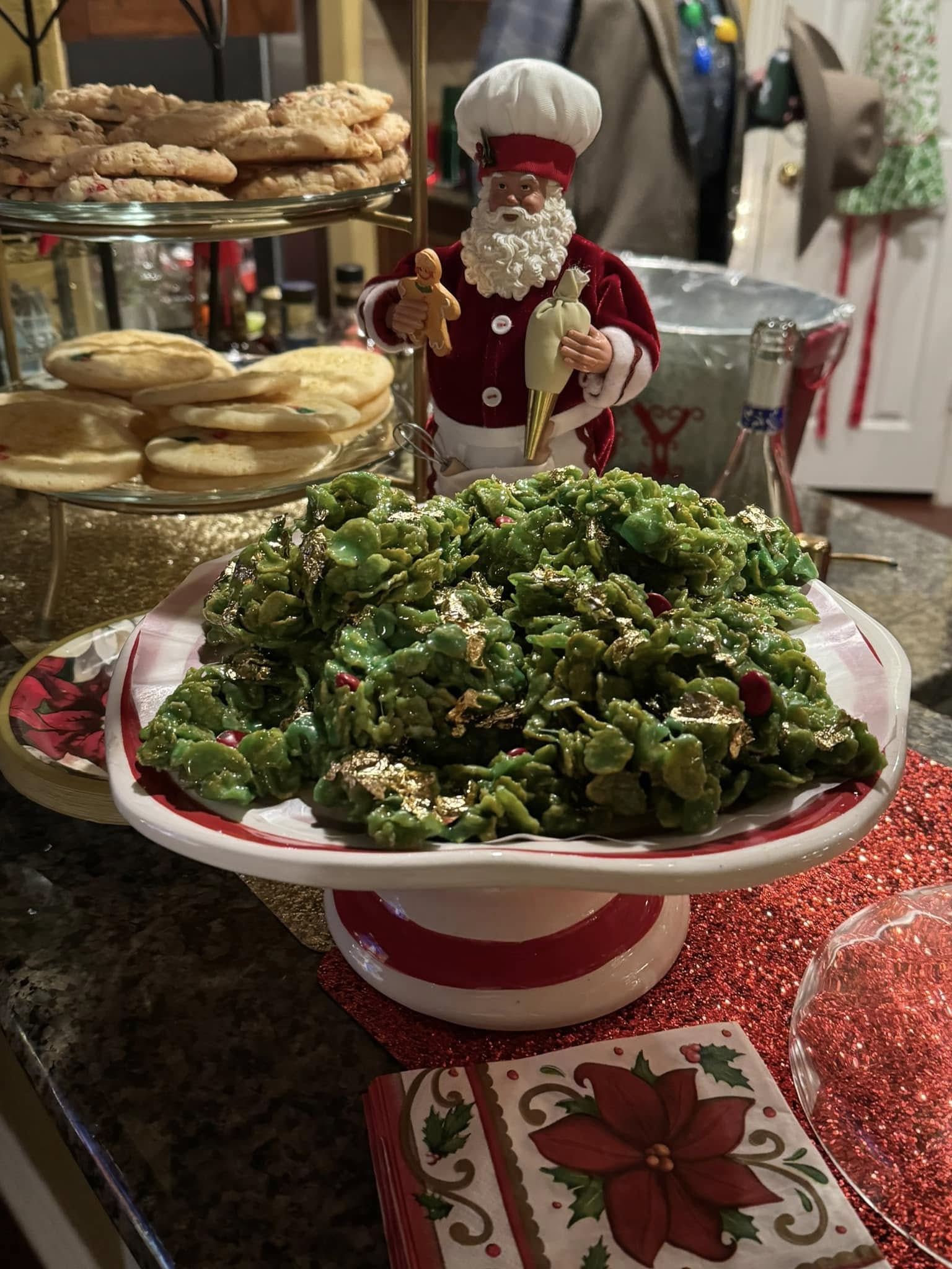 Green cornflake wreath cookies with red candies displayed on a cake stand next to a Santa Claus figure