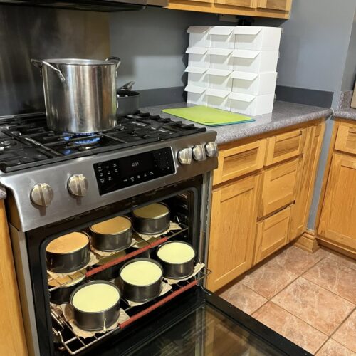 n open oven door showing two racks filled with 6-inch cheesecakes baking in water baths, with a large stockpot on the stove and stacks of white bakery boxes in the background