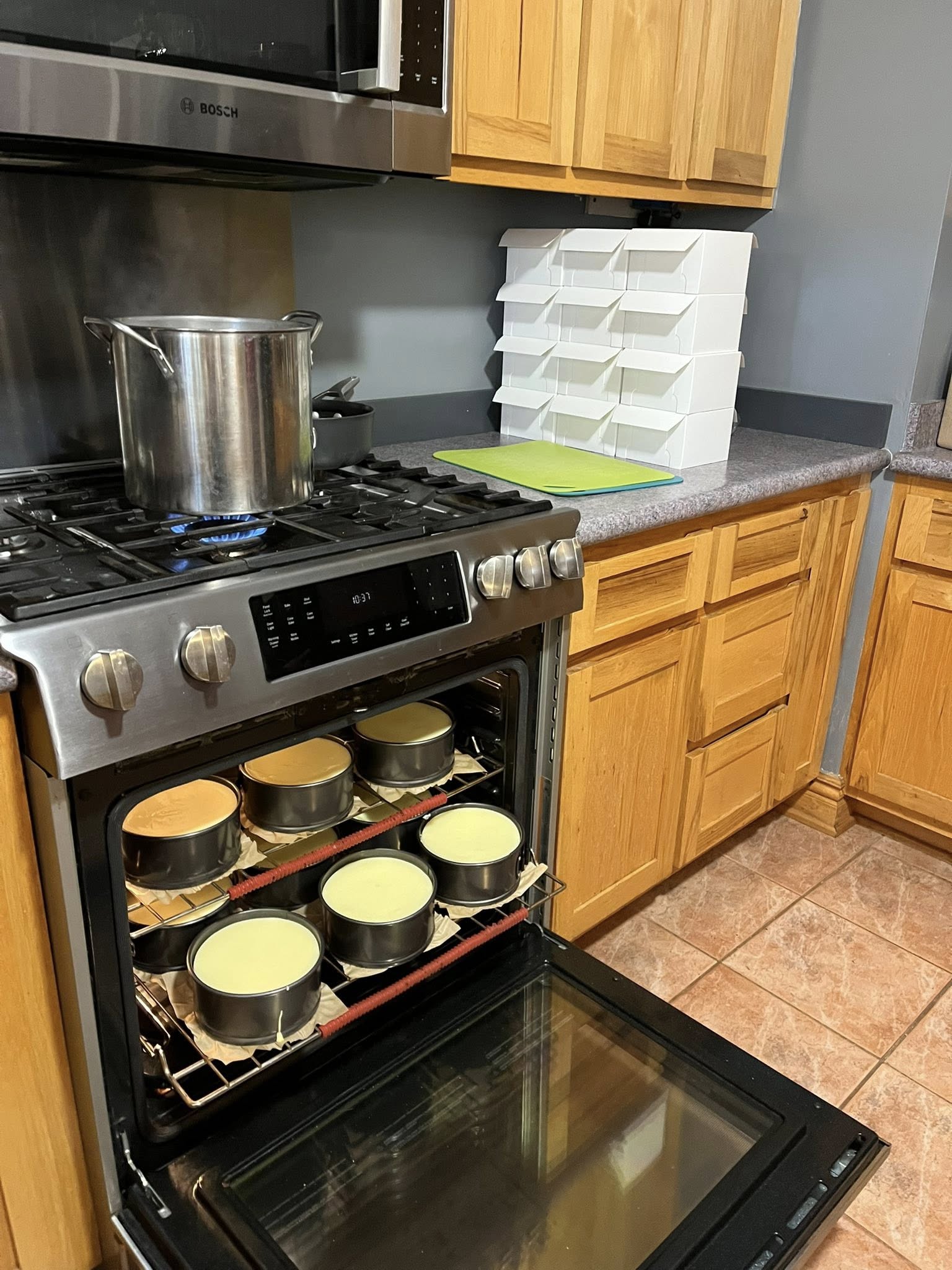 n open oven door showing two racks filled with 6-inch cheesecakes baking in water baths, with a large stockpot on the stove and stacks of white bakery boxes in the background