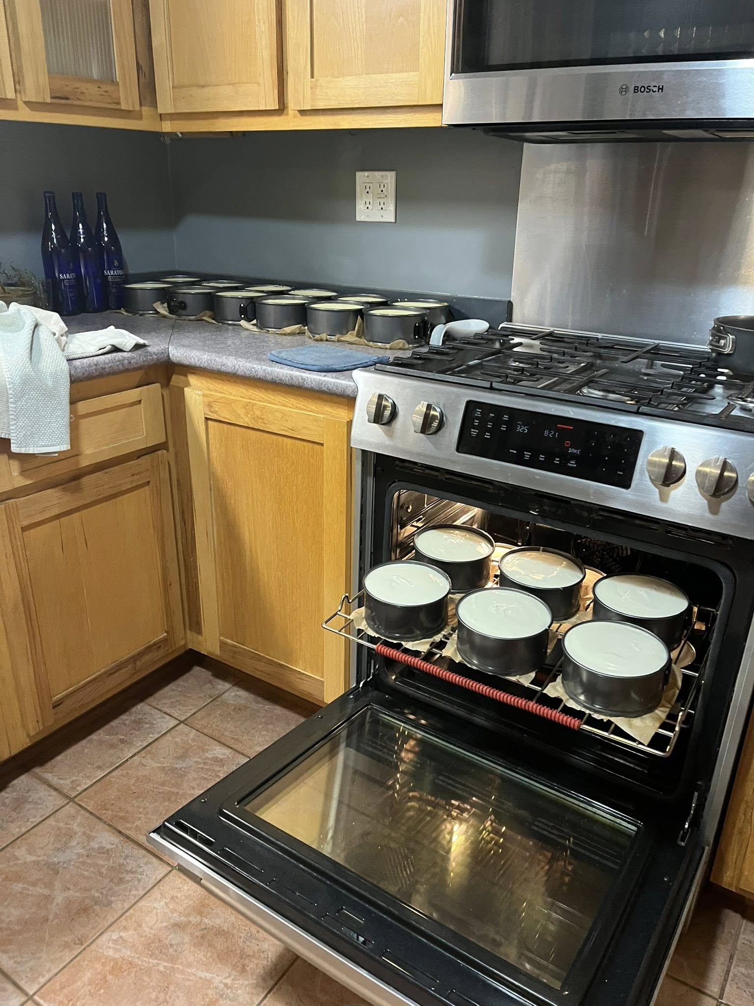 n open oven door showing two racks filled with 6-inch cheesecakes baking in water baths, with a large stockpot on the stove and stacks of white bakery boxes in the background