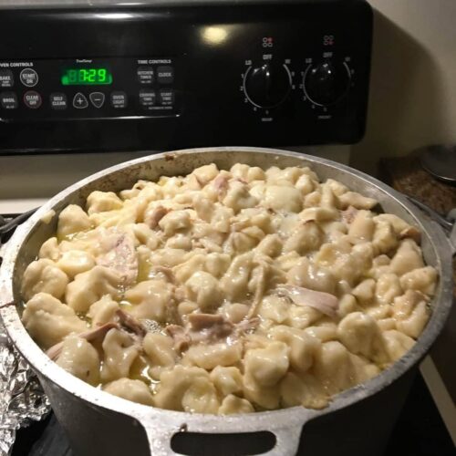 A large aluminum pot sitting on a stovetop, filled to the brim with rich chicken broth and plump, freshly cooked flat dumplings.