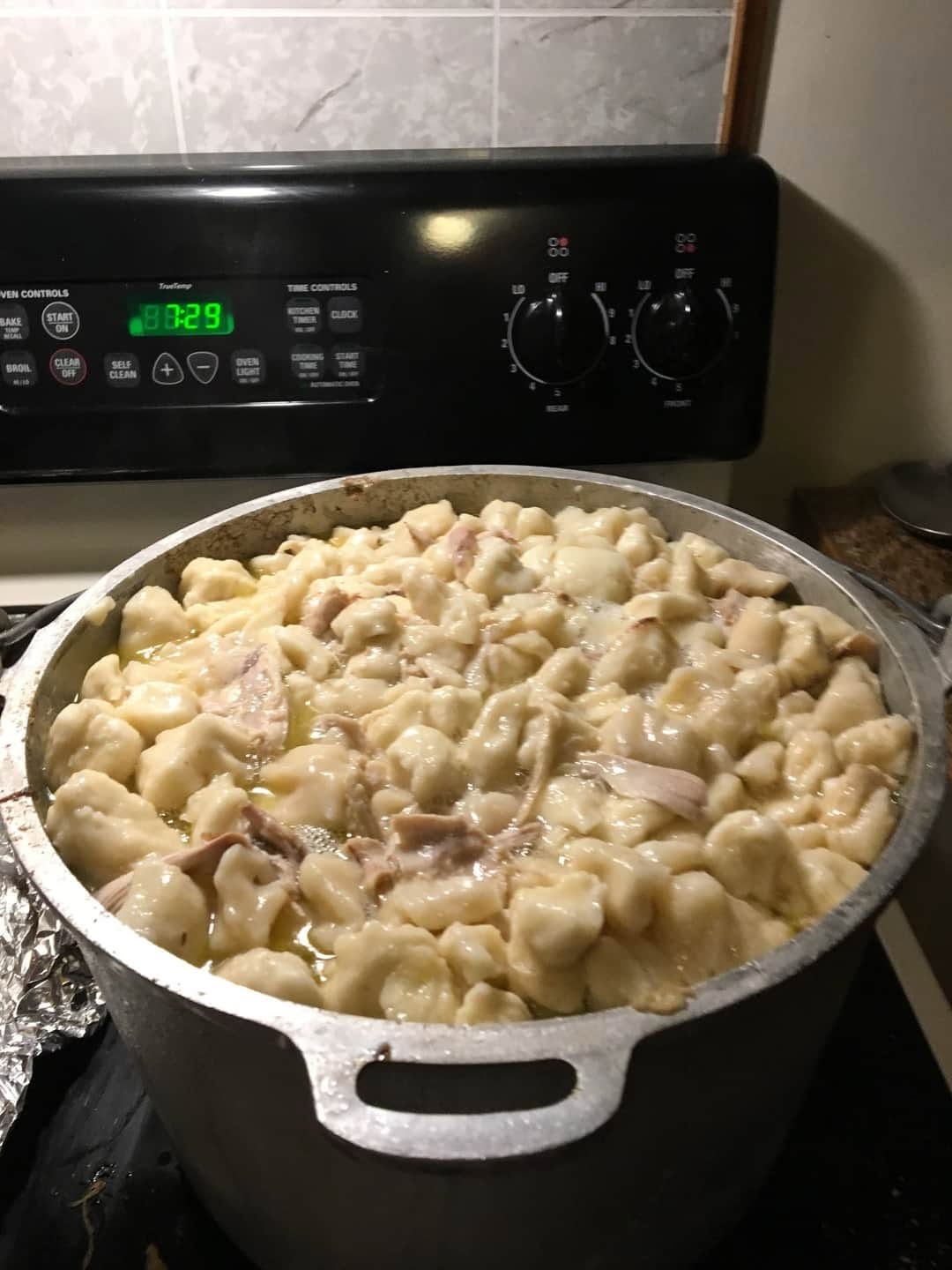 A large aluminum pot sitting on a stovetop, filled to the brim with rich chicken broth and plump, freshly cooked flat dumplings.