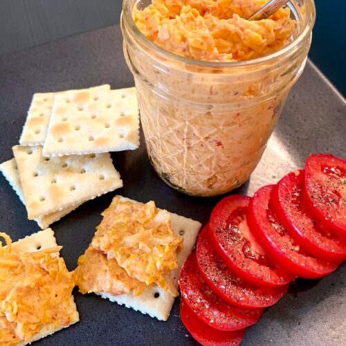 A glass jar filled with homemade homemade pimento cheese, served alongside saltine crackers and fresh sliced tomatoes with black pepper.