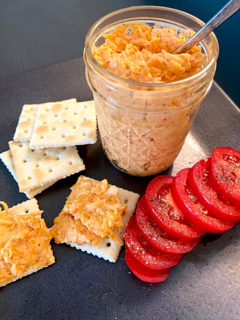 A glass jar filled with homemade homemade pimento cheese, served alongside saltine crackers and fresh sliced tomatoes with black pepper.