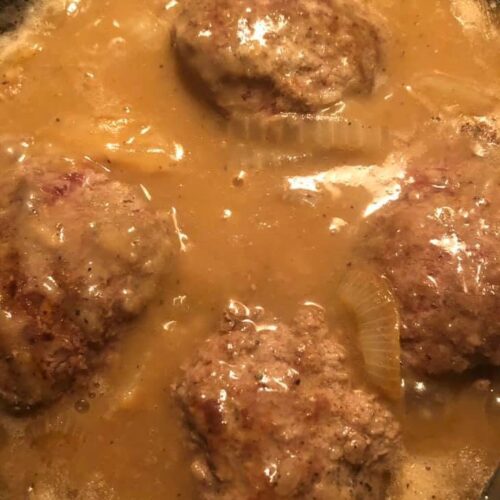 Four seasoned hamburger patties simmering in a rich, bubbling brown onion gravy in a cast iron skillet.