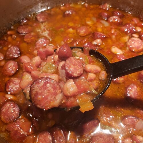 A black ladle lifting a hearty scoop of Southern pinto beans and sliced Conecuh smoked sausage from a simmering pot of rich, savory broth
