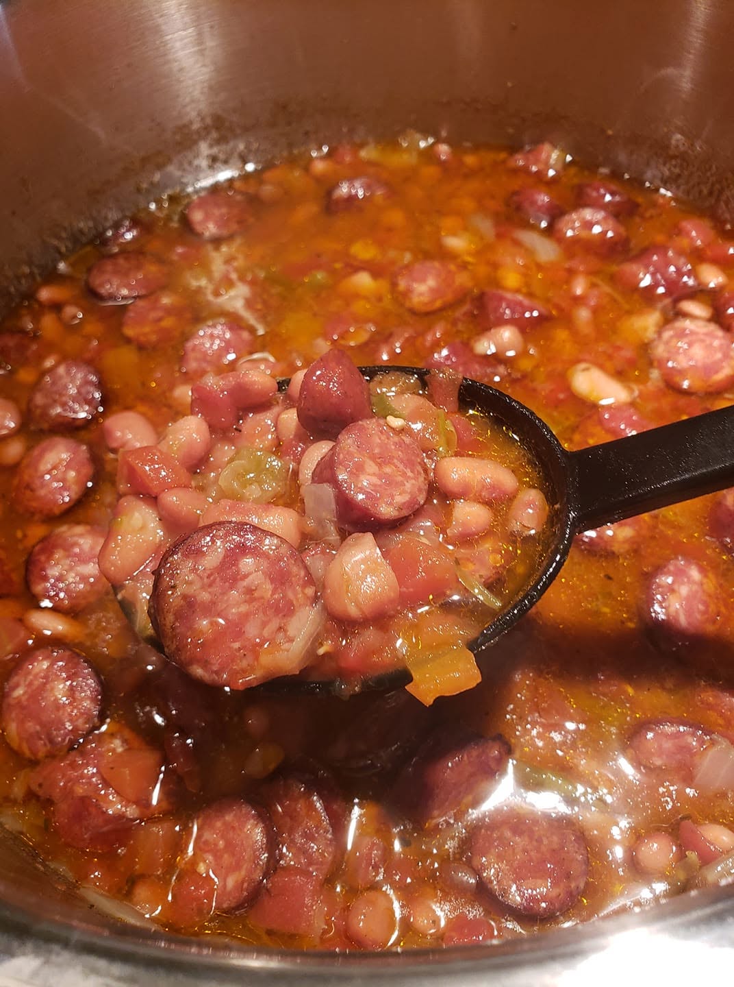 A black ladle lifting a hearty scoop of Southern pinto beans and sliced Conecuh smoked sausage from a simmering pot of rich, savory broth