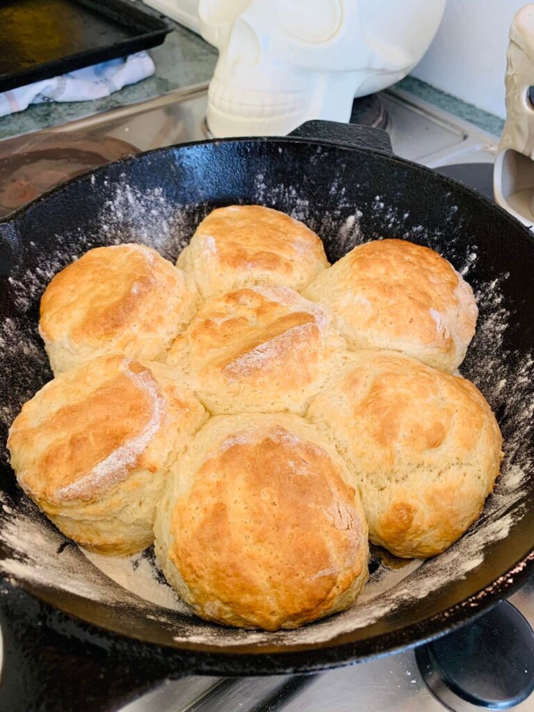 Seven large, golden-brown homemade buttermilk biscuits dusted with flour, baked together in a black cast iron skillet.