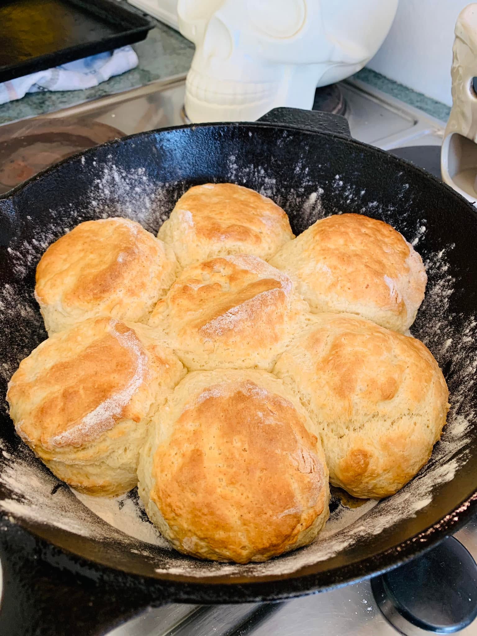Seven large, golden-brown homemade buttermilk biscuits dusted with flour, baked together in a black cast iron skillet.