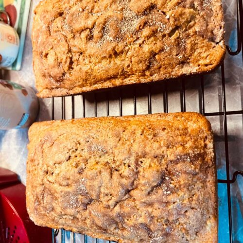 Two freshly baked loaves of golden apple pie bread topped with a sparkling cinnamon-sugar crust, resting on a black wire cooling rack.