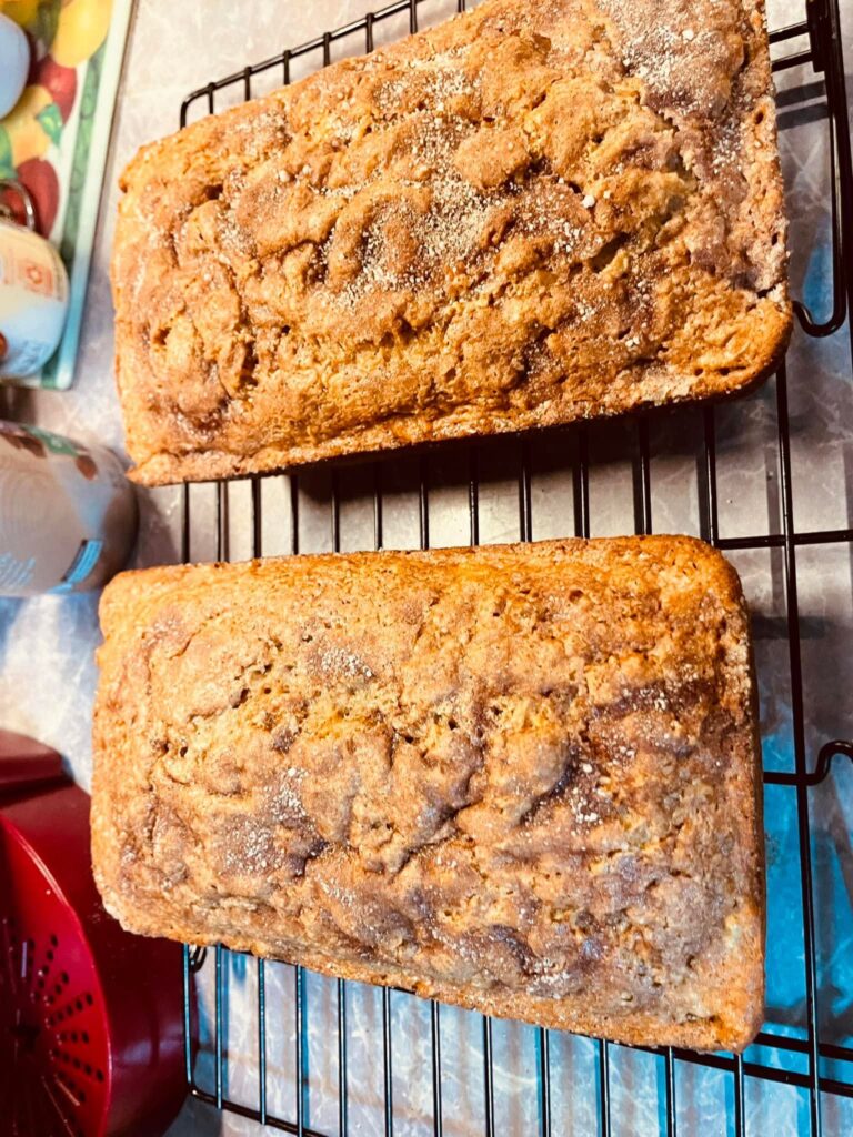 Two freshly baked loaves of golden apple pie bread topped with a sparkling cinnamon-sugar crust, resting on a black wire cooling rack.