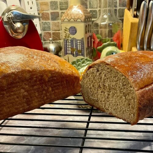 Two loaves of freshly baked apple butter whole wheat bread resting on a black wire cooling rack, with one loaf sliced to reveal a soft, dense, and hearty crumb.