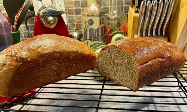Two loaves of freshly baked apple butter whole wheat bread resting on a black wire cooling rack, with one loaf sliced to reveal a soft, dense, and hearty crumb.