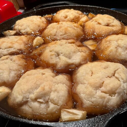 A cast iron skillet filled with golden brown, baked apple dumplings sitting in a rich, bubbling brown sugar syrup with chunks of apple scattered between them.