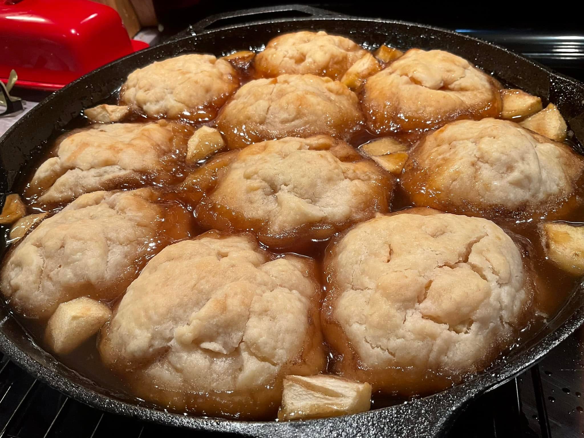 A cast iron skillet filled with golden brown, baked apple dumplings sitting in a rich, bubbling brown sugar syrup with chunks of apple scattered between them.