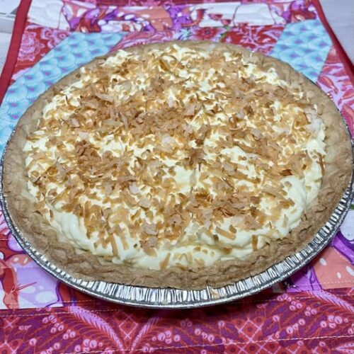 A beautiful homemade coconut cream pie in a foil pie tin, topped with a generous layer of toasted coconut flakes resting on a patterned pink quilt.