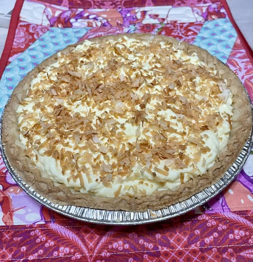 A beautiful homemade coconut cream pie in a foil pie tin, topped with a generous layer of toasted coconut flakes resting on a patterned pink quilt.