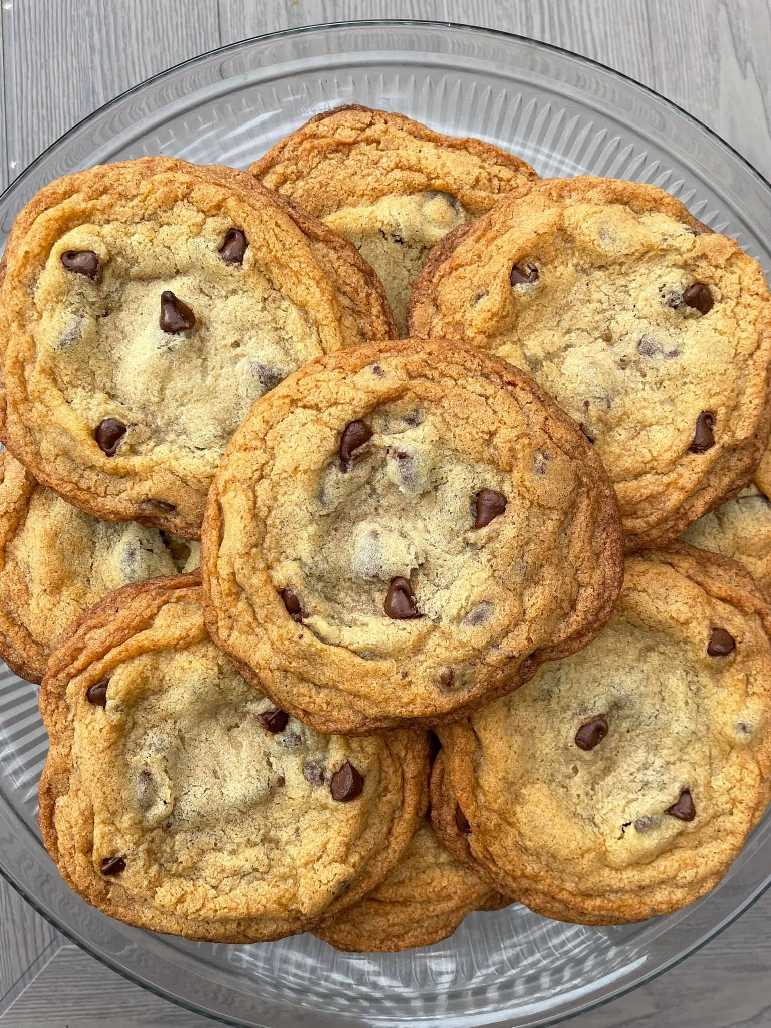 A clear glass platter overflowing with huge, golden-brown bakery style chocolate chip cookies featuring perfectly crisp edges and soft centers.
