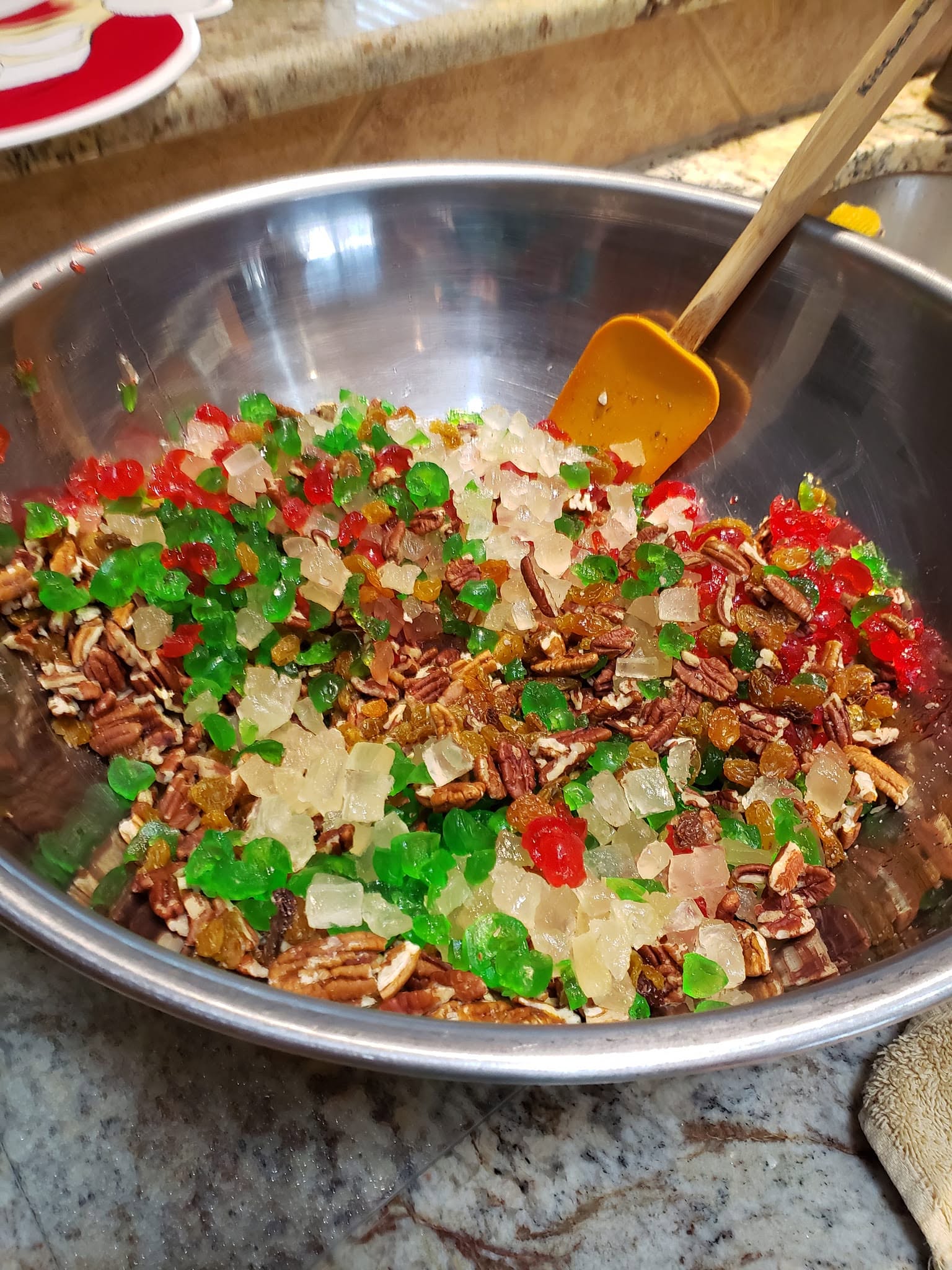 A massive stainless steel mixing bowl filled with chopped pecans, golden raisins, and vibrant red and green candied cherries and pineapple pieces.