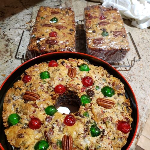 A beautifully baked, traditional Southern white fruitcake decorated with red and green candied cherries and pecan halves, surrounded by two smaller loaf pan fruitcakes.