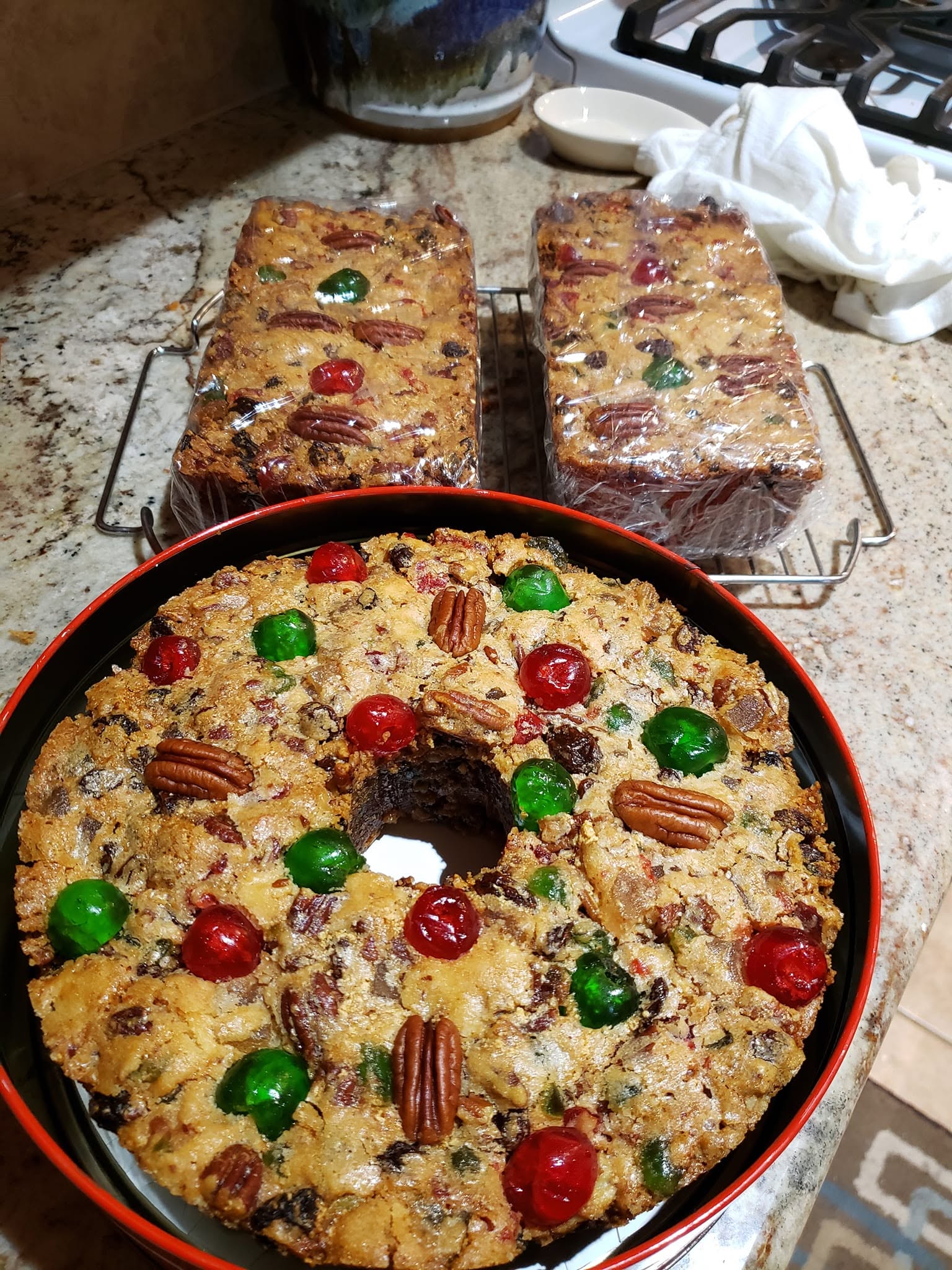 A beautifully baked, traditional Southern white fruitcake decorated with red and green candied cherries and pecan halves, surrounded by two smaller loaf pan fruitcakes.