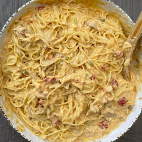 A large rustic bowl overflowing with creamy, cheesy chicken spaghetti speckled with diced tomatoes and green peppers.