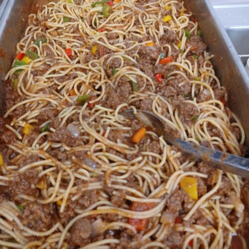 A massive stainless steel catering pan filled to the brim with spaghetti, ground beef, and colorful bell peppers tossed in a rich tomato sauce.