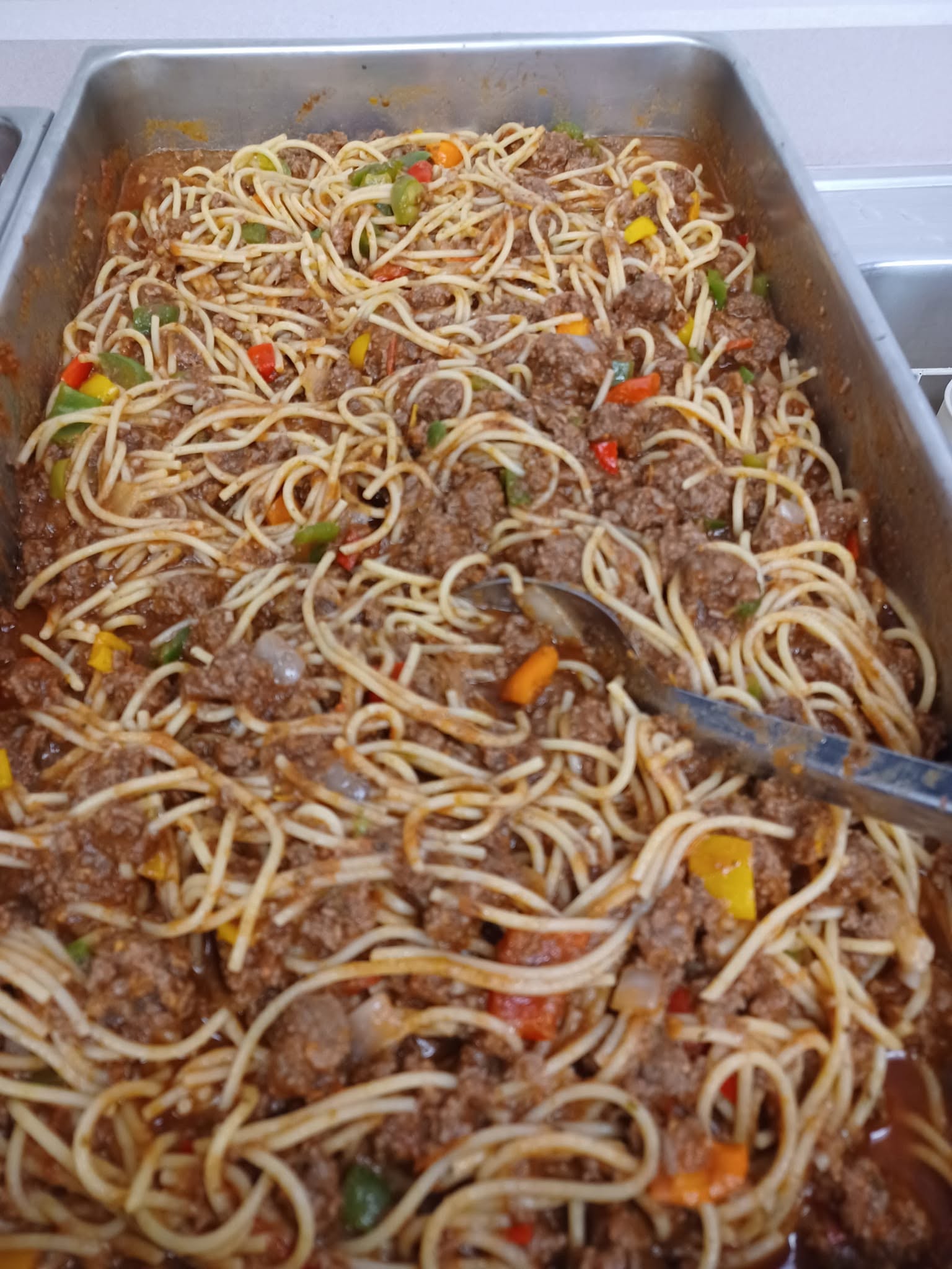 A massive stainless steel catering pan filled to the brim with spaghetti, ground beef, and colorful bell peppers tossed in a rich tomato sauce.