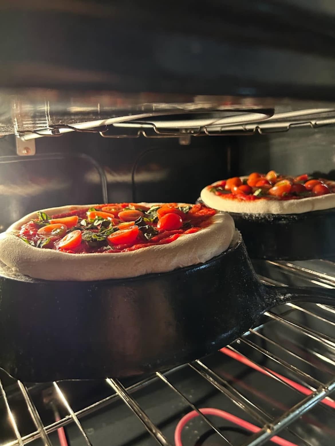 Two personal-sized mini pizzas topped with red sauce, fresh cherry tomatoes, and green basil leaves, baking directly on the flat bottoms of upside-down vintage cast iron skillets inside a hot oven.