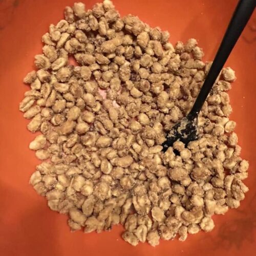 A bright orange bowl filled with a fresh batch of candied maple peanuts and a black silicone spatula resting inside.