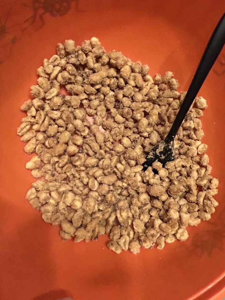 A bright orange bowl filled with a fresh batch of candied maple peanuts and a black silicone spatula resting inside.