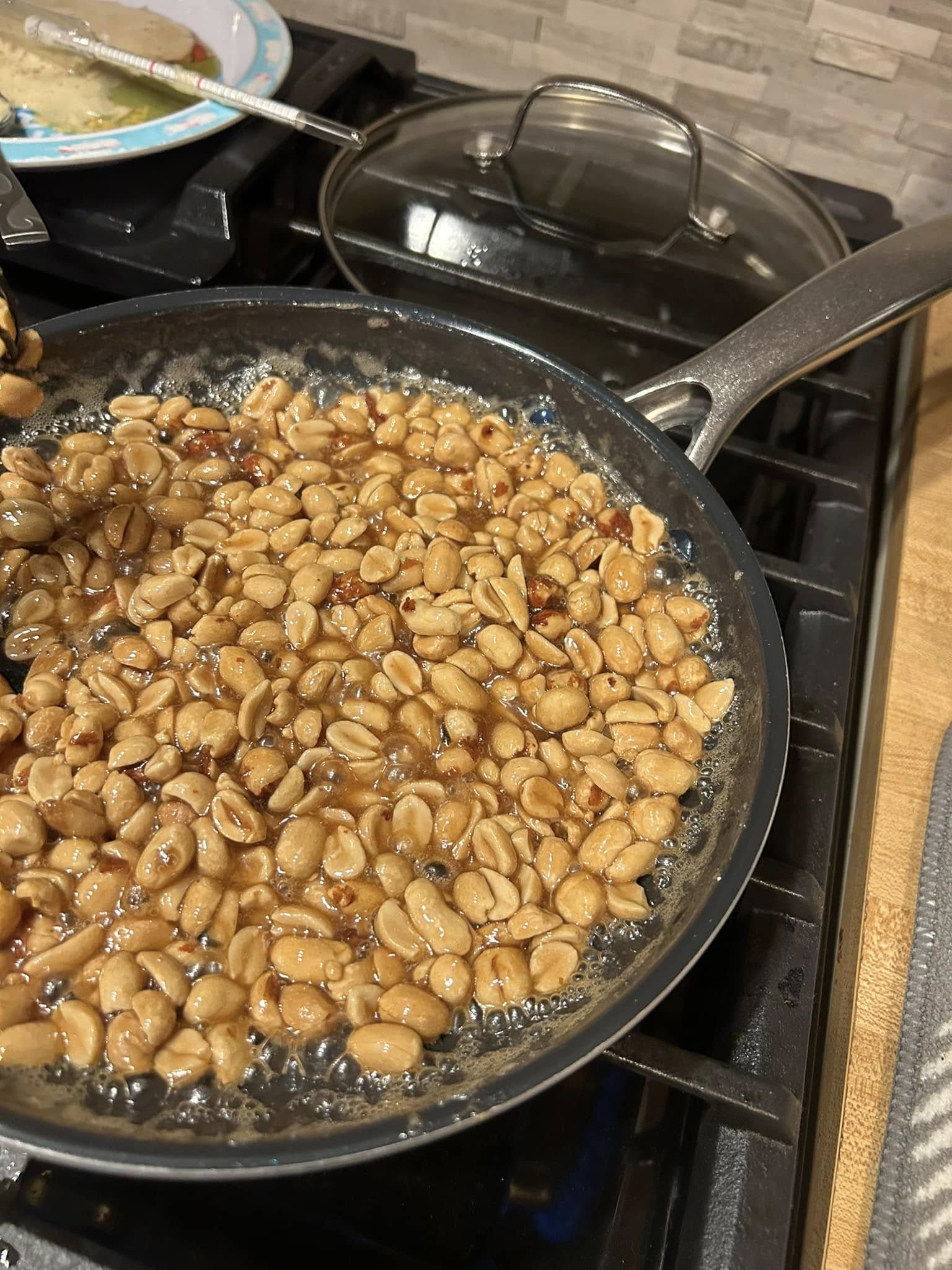 A black whisk continuously stirring a bubbling mixture of peanuts and thickening maple syrup in a nonstick skillet.