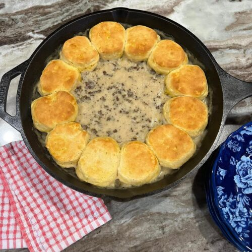 A black cast iron skillet filled with creamy sausage gravy in the center, surrounded by a ring of perfectly golden-brown baked biscuits.