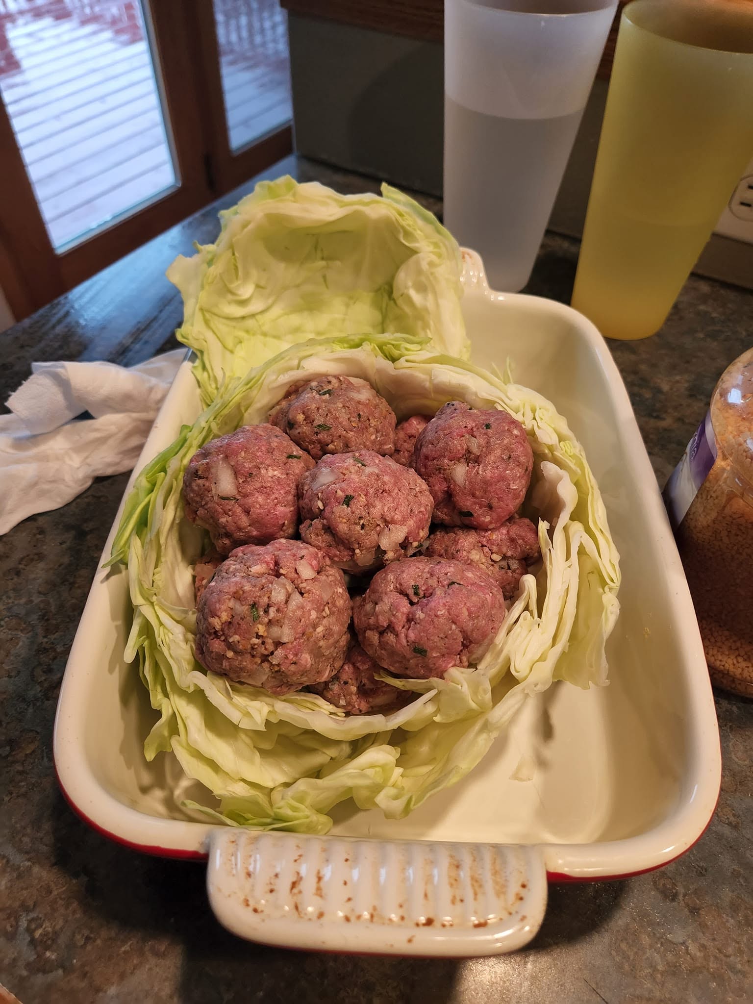 A hollowed-out green cabbage bowl sitting in a white baking dish, packed full of large, raw, seasoned meatballs.